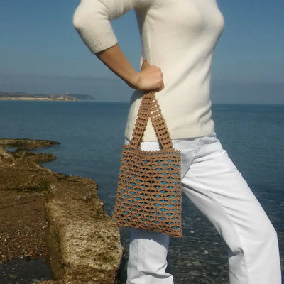 Handmade brown crochet tote bag with geometric mesh pattern, short handles, displayed by a person near water.