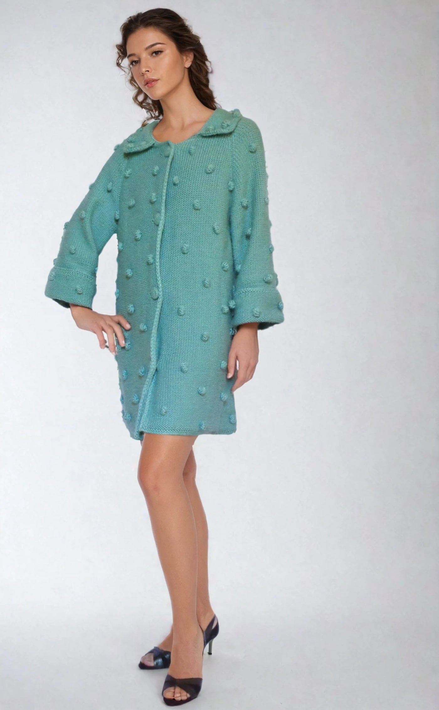 Woman wearing a handmade turquoise knitted coat with bubble texture and oversized sleeves, styled in a cozy, vintage-inspired look against a neutral background.