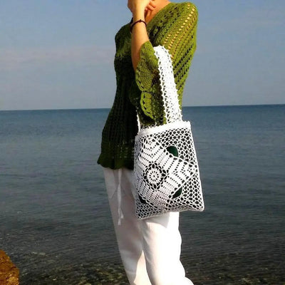 White crochet tote bag with openwork design and two-sided handles, paired with a green linen top by the sea.