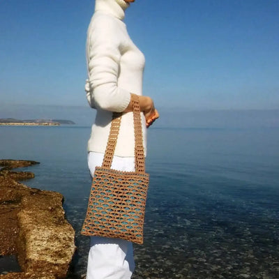 Handmade small brown crochet tote bag with geometric mesh pattern, featuring short handles and blue lining, standing by the sea.